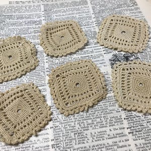 May include: Five square beige crochet coasters featuring a central hole and intricate openwork patterns. Each coaster has a scalloped edge and is crafted from fine thread, displaying a consistent geometric design across all five individual pieces.