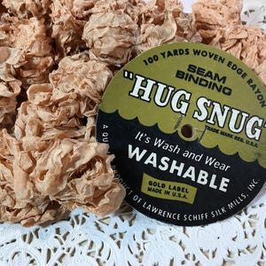 May include: Close-up of a pile of peach-colored woven edge rayon seam binding. A circular label reads "HUG SNUG" and "WASHABLE". The label also states "100 yards" and "Made in U.S.A."