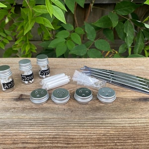 May include: A selection of small glass bottles with silver lids, each labelled with a different spice name, such as "Birch," "Anis," "Clove," and "Cyp." Also includes small metal containers, tweezers, and other small items, all arranged on a wooden surface.
