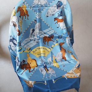 May include: A light blue silk scarf with a horse-themed design. The scarf features a repeating pattern of horses, riders, and the text "Musée Vivant du Cheval Chantilly". The scarf is draped over a white chair.