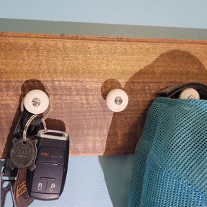 May include: A wooden wall-mounted key rack with two white ceramic hooks. A blue mesh hat hangs on one hook and a set of keys with a car key and a round key fob hang on the other hook.