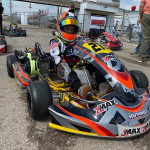 Peut inclure: Une personne portant une combinaison et un casque de course est assise dans un karting. Le karting est noir, gris et orange avec le numéro 21. Le casque a une visière miroir. Le karting porte le texte "ZMAX" et "SOUTHWEST REGIONAL CUP".