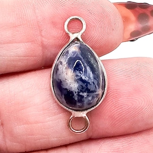 May include: A teardrop-shaped pendant featuring a polished, deep blue and white mottled stone set in a silver-toned metal frame. The pendant includes a small circular jump ring at both the top and the bottom for attachment to jewelry chains.