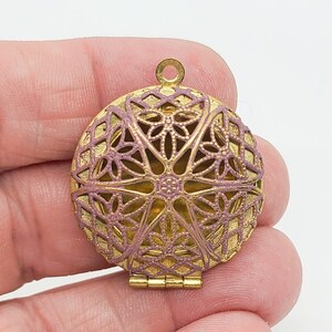 Peut inclure: Un médaillon rond en métal doré présentant un motif complexe en filigrane ajouré sur le couvercle avant. Le motif en relief et texturé est rehaussé d'une finition mauve sourd ou violet poussiéreux, avec une petite bélière circulaire au sommet.