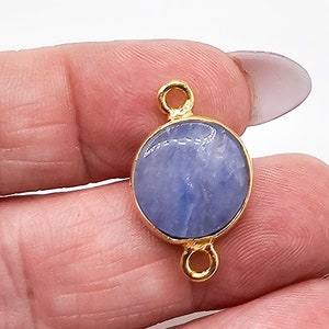 May include: A circular pendant featuring a smooth, light blue stone set within a polished gold-toned metal frame. The piece includes two small, circular gold-toned loops, one positioned at the top and one at the bottom, for attachment purposes.