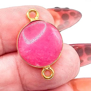 May include: A circular jewelry connector featuring a vibrant hot pink stone set within a polished gold-toned metal frame. The piece includes two small, circular gold-toned loops positioned at the top and bottom for attachment purposes.