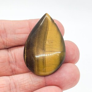 Peut inclure: Une pierre précieuse œil-de-tigre polie en forme de goutte, dotée d'une surface lisse et réfléchissante avec des bandes alternées de fibres chatoyantes jaune doré et brun foncé créant un effet scintillant sur la face incurvée.