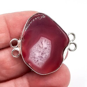 Peut inclure: Une tranche d'agate magenta de forme irrégulière polie, sertie dans une monture en métal argenté. Le centre présente une inclusion de druse blanche cristalline et texturée. La pièce comporte deux petites boucles en métal argenté de chaque côté.