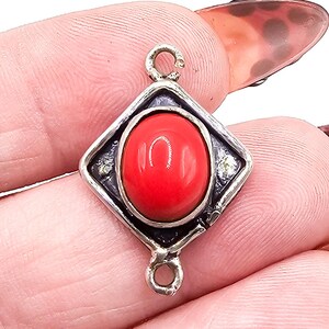 May include: A diamond-shaped silver pendant featuring a smooth, oval-cut red stone set in the center. The metal frame has a dark, oxidized finish and includes two small circular loops at the top and bottom for attachment to a chain or cord.