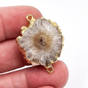May include: A circular, translucent white and light brown agate slice pendant featuring a textured, crystalline center and a jagged, natural edge. The stone is encased in a bright gold-toned metal border and includes two small gold-toned metal loops.