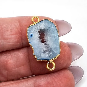 May include: A light blue druzy agate gemstone connector with a jagged, crystalline center and a gold-toned metal rim. The connector features two small, circular gold-toned metal loops attached at the top and bottom for jewelry assembly.