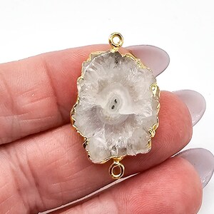 May include: A translucent white druzy quartz gemstone connector featuring a natural, irregular shape with a textured, crystalline surface. The stone is encased in a thin, polished gold-tone metal rim and includes two small circular loops at the top and bottom.