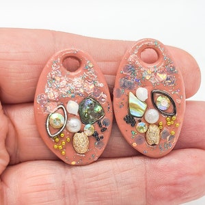 May include: Two oval-shaped terracotta-colored pendants featuring a hole at the top. Each piece is decorated with iridescent glitter, small white pearls, gold-toned beads, metallic accents, and fragments of iridescent abalone shell inlay.