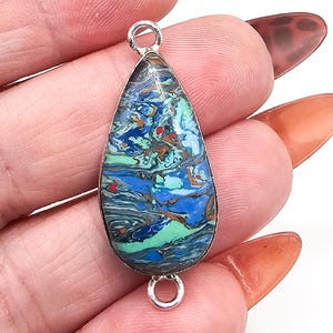 May include: A teardrop-shaped pendant featuring a swirling marbled pattern of blue, green, and brown tones encased in a silver-toned metal frame. The pendant includes a small circular jump ring at both the top and bottom for attachment purposes.