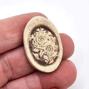 May include: An oval-shaped cameo brooch featuring a raised, cream-colored floral relief design set against a dark brown, recessed background. The piece has a textured, matte finish and a smooth, rounded border surrounding the central flower motif.