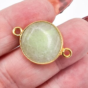 May include: A round, pale green gemstone connector featuring a smooth, polished surface set within a thin gold-toned metal bezel. The connector includes two small, circular metal loops extending from opposite sides of the central stone setting.