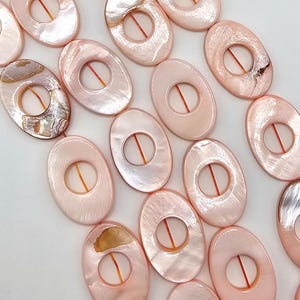 Peut inclure: Une collection de perles ovales en nacre rose clair, chacune dotée d'une découpe circulaire centrale. Les perles sont enfilées sur un fin cordon orange, présentant une surface lisse et irisée avec des variations naturelles de texture et de couleur.