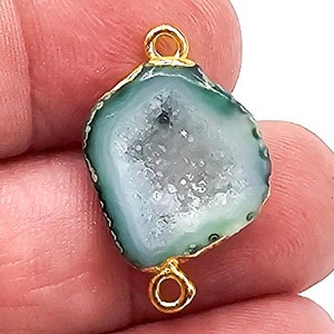 May include: A small, irregular-shaped teal agate druzy connector featuring a sparkling, crystalline center and a smooth, polished outer edge. The stone is encased in a thin gold-toned metal border and includes two small gold-toned metal loops at the top and bottom for attachment.
