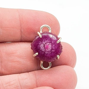 May include: A circular, deep magenta druzy stone connector featuring a textured, crystalline surface. The stone is secured by four silver-toned metal prongs and includes two small, circular silver-toned metal loops at the top and bottom.