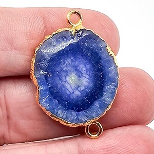 May include: A circular blue druzy gemstone connector featuring a textured, crystalline surface and a vibrant deep blue hue. The stone is encased in a metallic gold-toned rim and includes two small gold-toned circular loops at the top and bottom.