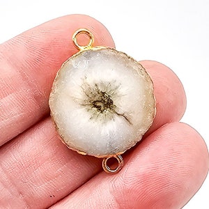 May include: A circular white druzy stone connector featuring a textured, crystalline surface with a dark center. The stone is encased in a gold-toned metal rim and includes two small gold-toned metal loops at the top and bottom for attachment.