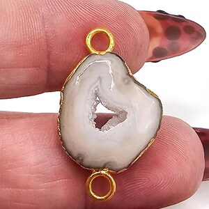 May include: A white agate gemstone connector featuring a central geode cavity and a gold-toned metal rim. The piece includes two small circular gold-toned metal loops, one at the top and one at the bottom, for attachment purposes.