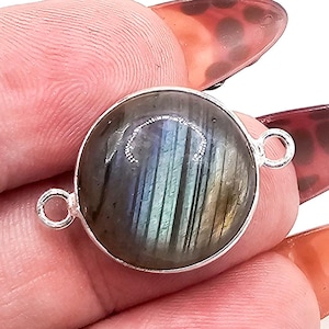 Op de afbeelding: Een ronde labradoriet edelsteen connector met een gepolijst oppervlak met iriserende blauwe en grijze flitsen, gevat in een eenvoudige zilverkleurige metalen zetting met twee kleine ronde lusjes aan de tegenoverliggende zijden.