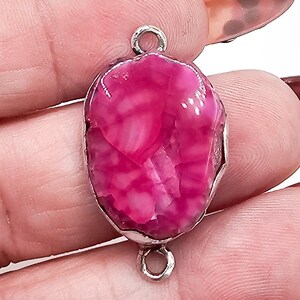 May include: A vibrant hot pink oval gemstone connector featuring a polished, faceted surface with natural crackled patterns. The stone is encased in a silver-toned metal setting with a small circular loop at both the top and the bottom for attachment.