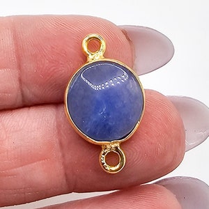 May include: A circular blue gemstone connector featuring a smooth, polished surface set within a gold-toned metal bezel. The connector includes two small, circular loops at the top and bottom, designed for attachment to jewelry chains or findings.