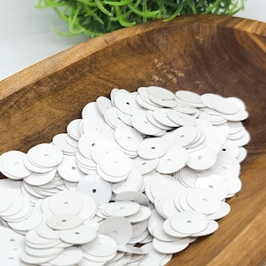 May include: A collection of small, circular, matte white sequins, each featuring a single centered hole for attachment. These flat, round embellishments are presented in a smooth, dark brown wooden bowl with a visible natural wood grain.