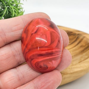 Peut inclure: Un cabochon de forme ovale présentant une surface lisse et polie avec un motif tourbillonnant de tons rouge profond, rouge cramoisi vif et noir foncé. La pièce a une finition brillante qui reflète la lumière, créant un aspect marbré vibrant.