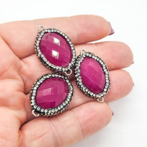 May include: Three oval-shaped magenta jewelry connectors featuring faceted surfaces. Each piece is framed by a dark metal border encrusted with small, clear, sparkling rhinestones. Each connector includes two small metal loops for attachment.