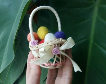 May include: A small wicker basket with a white handle and a white ribbon is filled with colorful felt balls. The basket is decorated with pink felt flowers.