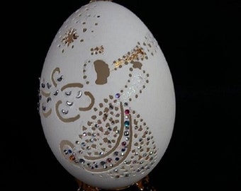 Puede incluir: Un huevo blanco con una talla detallada de un ángel, decorado con diamantes de imitación y detalles dorados. El huevo se muestra en un soporte dorado.