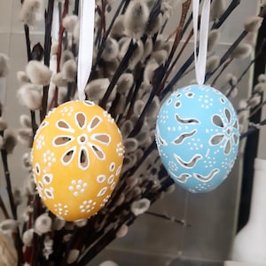 Puede incluir: Dos adornos decorativos de huevos de Pascua. Uno es amarillo con recortes florales blancos y detalles de cuentas blancas. El otro es azul claro con recortes de pájaros y flores blancas. Ambos cuelgan de cintas blancas sobre un fondo de ramas de sauce.