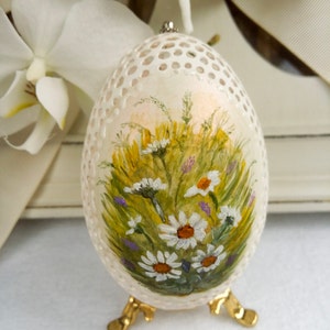 May include: An intricately carved eggshell with a painted floral design. The egg features a field of daisies and other wildflowers in shades of white, yellow, green, and purple. It rests on a gold-colored metal stand, showcasing the delicate artwork.