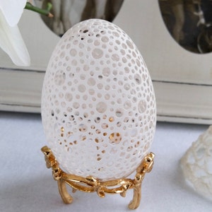 May include: A white egg with a delicate, intricate lace-like pattern, displayed on a gold metal stand.