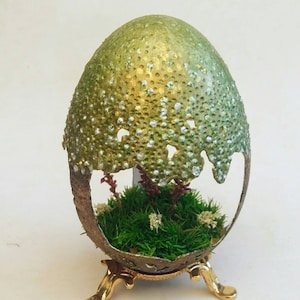 Pode incluir: Um ovo decorativo com um acabamento salpicado dourado e verde, com uma pequena abertura esculpida que revela um jardim de musgo em miniatura no seu interior. O ovo está empoleirado em um suporte dourado com três pernas.