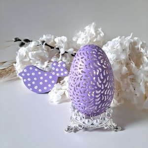 Peut inclure: Un œuf de Pâques violet, sculpté de manière complexe, avec une figurine d'oiseau à pois blancs. L'œuf est sur un support argenté avec des accents de perles blanches. L'œuf et l'oiseau sont entourés de fleurs blanches.