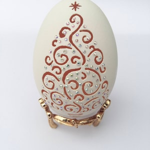 May include: A cream-colored egg-shaped ornament with a brown, swirling tree design and sparkling crystal accents. The ornament sits on a gold-colored, ornate stand. A star is at the top of the tree design.