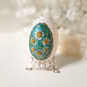 May include: A delicate, hand-carved egg with a teal blue background and white flowers painted on it. The egg is displayed on a silver stand with white pearls.