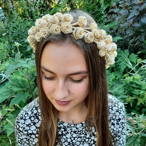 Könnte beinhalten: Ein floraler Kranz aus beigefarbenen Papierrosen. Der Kranz wird auf dem Kopf einer Person getragen, mit langen braunen Haaren und einem Oberteil mit schwarz-weißem Blumenmuster. Der Hintergrund ist ein üppiger grüner Garten.