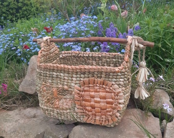 May include: A woven basket with a natural brown and tan color scheme. The basket has a wooden handle and two tassels. The basket is sitting on a stone wall in a garden setting.