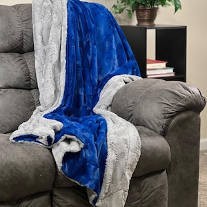 May include: A dual-sided throw blanket featuring a vibrant royal blue side with a textured, swirling pattern and a contrasting light gray side with a soft, plush, rippled texture, draped over a dark gray upholstered sofa cushion.