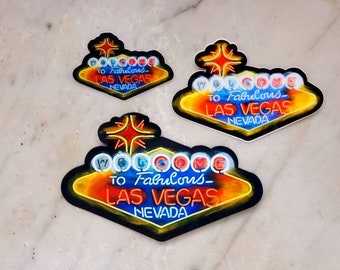Könnte beinhalten: Drei verschiedene Größen von Aufklebern mit der Aufschrift "Willkommen im fabelhaften Las Vegas, Nevada". Die Aufkleber haben die Form des ikonischen Las Vegas-Schilds mit einem roten Stern oben. Das Schild ist schwarz umrandet und hat einen gelben, orangen und roten Neon-Effekt. Der Text ist weiß.