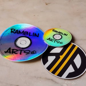 Puede incluir: Tres pegatinas redondas con un acabado holográfico iridiscente. La pegatina más grande presenta el texto "Ramblin Artso" en letras negras. La pegatina más pequeña presenta el mismo texto en un degradado arcoíris. La tercera pegatina es negra con un círculo blanco y dos rayas amarillas.
