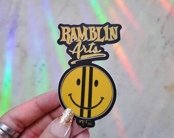 May include: A yellow smiley face sticker with black stripes and a black outline. The sticker says "Ramblin Arts" in black and yellow lettering.