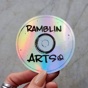 Puede incluir: Una pegatina holográfica con el texto "Ramblin Artso" en letras negras con un signo de paz. La pegatina es redonda y tiene un fondo iridiscente arcoíris.