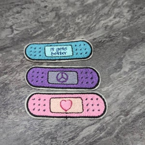 May include: Three embroidered iron-on patches shaped like plasters. The first is light blue with the text "It gets better". The second is purple with a peace sign. The third is pink with a heart.