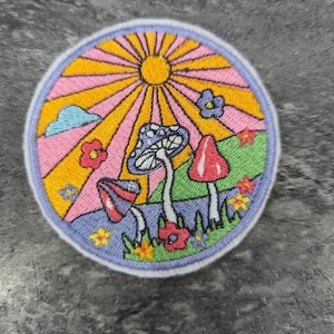 May include: A circular embroidered patch featuring a psychedelic sun, clouds, flowers, and mushrooms. The sun is yellow with orange rays, the clouds are light blue, the flowers are pink, yellow, and blue, and the mushrooms are red, white, and purple.